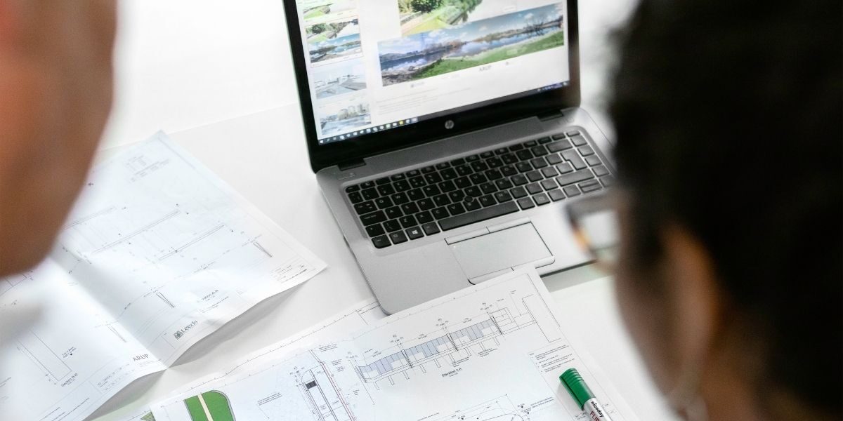 Image of a man and a woman looking at a digital representation of an engineering plan. Paper engineering plans on the table in front of them.