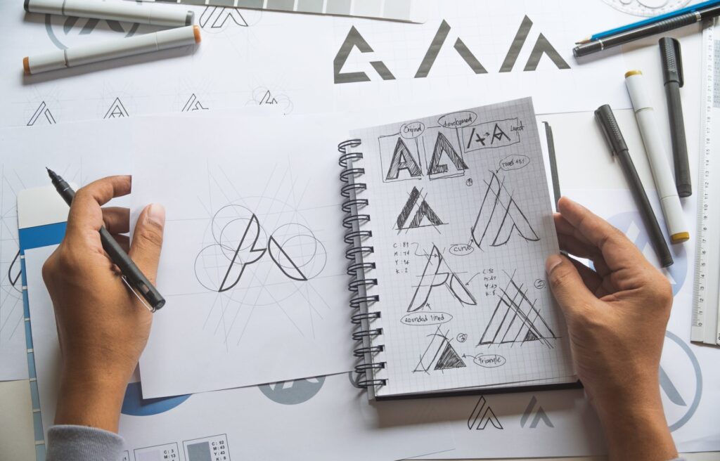 Top down view of a desk with a designer working on logo designs, stationary is lightly scattered on the desk and different print outs of logo variations are overlapping eachother.