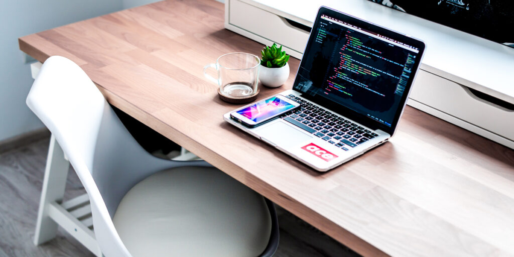 A laptop sits on a wooden desk with a phone resting on top of it.