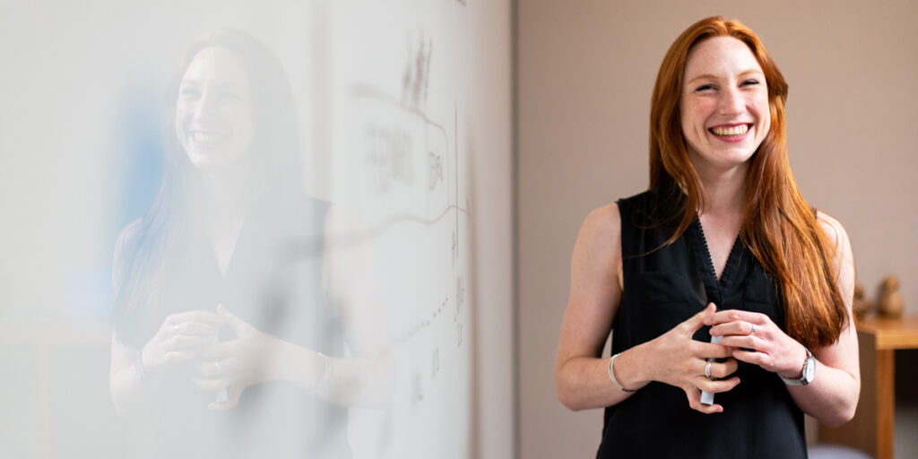 Person with long red hair stands smiling in front of a white board.