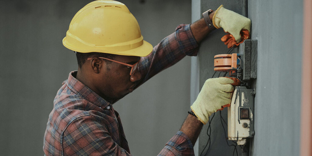 A man (Gas Engineering Operative) in a yellow helmet wiring an electrical appliance on a grey wall