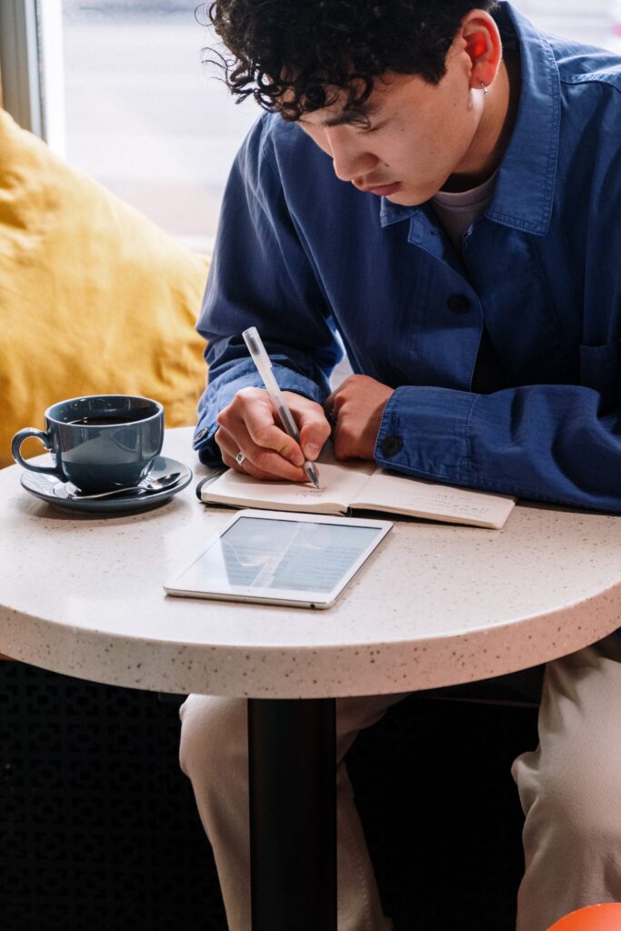 A man sitting at a coffee shop enjoying a cup of coffee whilst planning on a notebook and looking at a iPad.