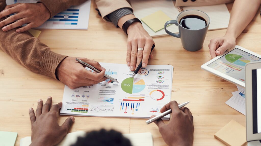 A Digital Marketing team sitting around a table with coffee, notebooks and charts deciding on analytics and pointing at a pie chart.