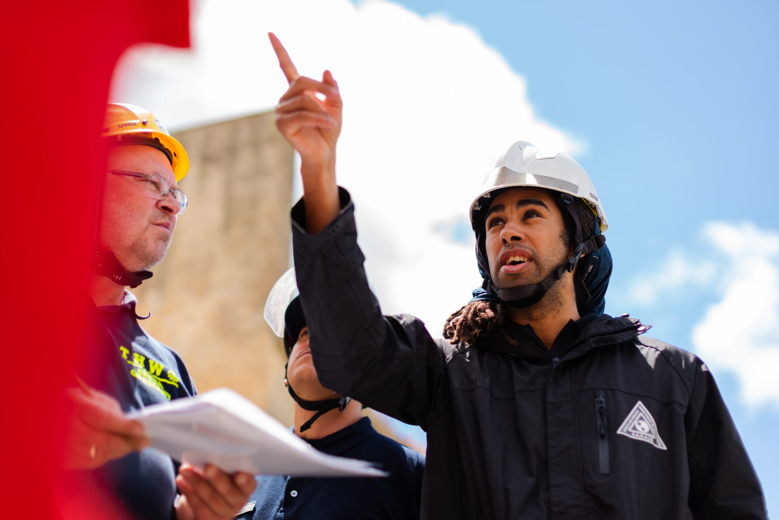 A man, water technician, in a white helmet points to something, while another man in a yellow helmet is holding papers.