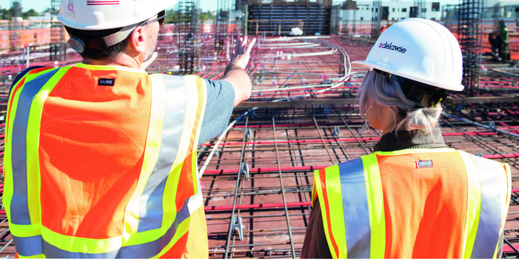 Two construction workers wearing helmets and high visibility jackets are looking out towards a building site.