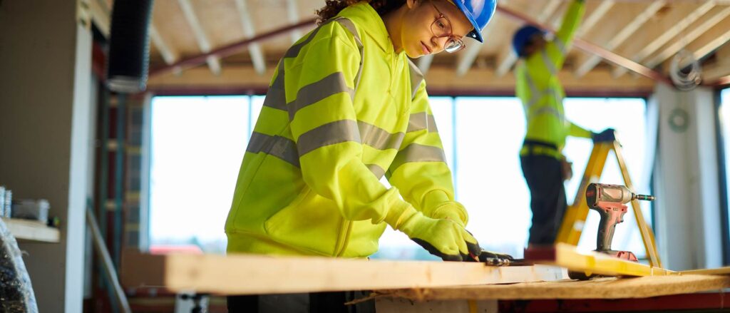 Feature a young girl, a level 2 carpentry and joinery apprentice, measuring and working with wood on a crafting table.