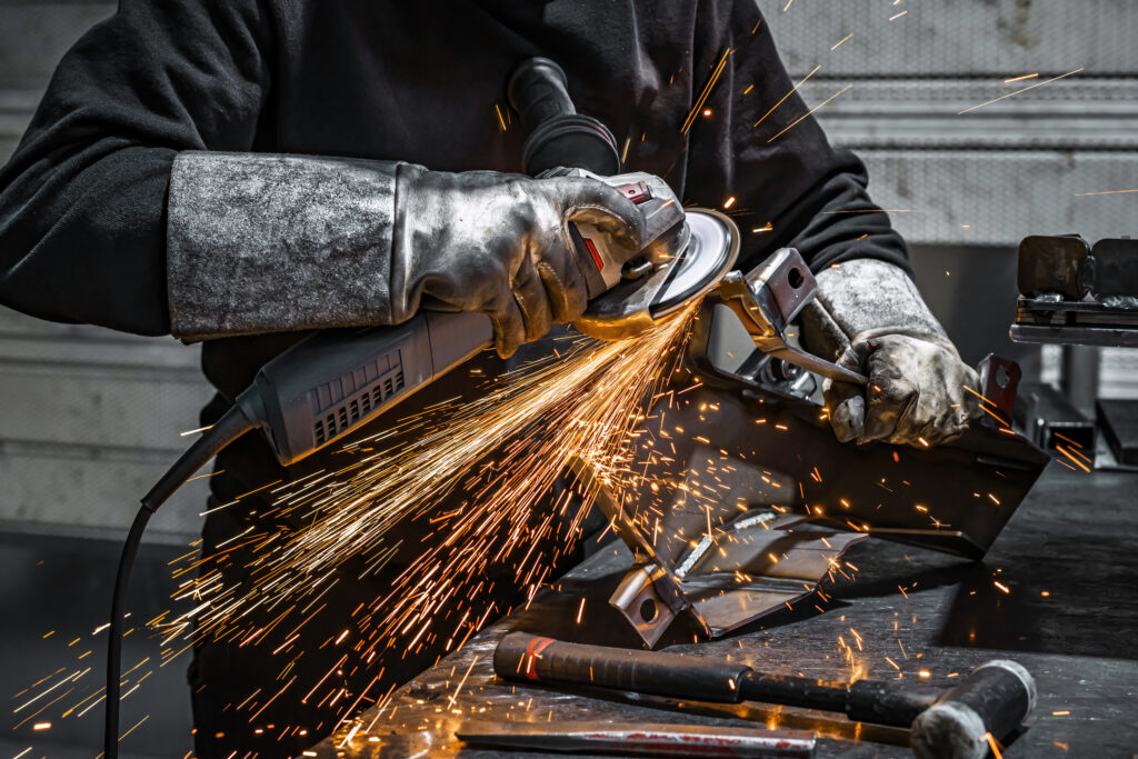 A view of the silver gloved hands of a worker grinding a piece of metal causing sparks to ignite